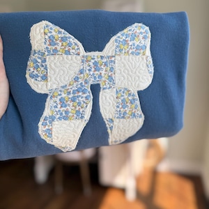 May include: A blue sweatshirt featuring a large, centered bow applique. The bow is constructed from alternating patches of a floral fabric with blue, yellow, and white blossoms and a solid cream-colored fabric with a quilted, wavy stitch pattern.