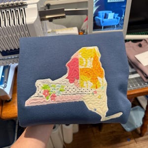 K&ouml;nnte beinhalten: Ein marineblaues Sweatshirt mit einem gesteppten Design des Umrisses des Bundesstaates New York. Das Design beinhaltet florale, Blatt- und Punktmuster in Rosa, Gelb und Wei&szlig;. Das Sweatshirt ist gefaltet und zeigt die detaillierte Stickerei.