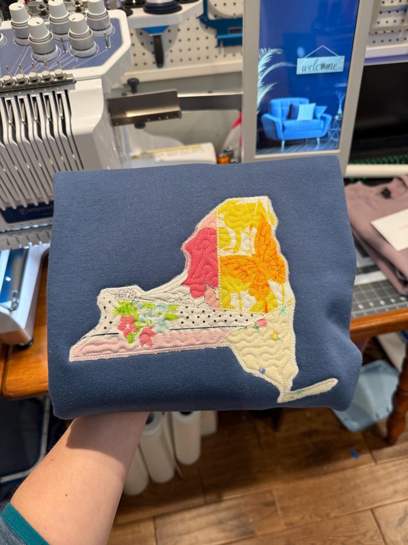 K&ouml;nnte beinhalten: Ein marineblaues Sweatshirt mit einem gesteppten Design des Umrisses des Bundesstaates New York. Das Design beinhaltet florale, Blatt- und Punktmuster in Rosa, Gelb und Wei&szlig;. Das Sweatshirt ist gefaltet und zeigt die detaillierte Stickerei.