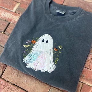 May include: A charcoal gray crewneck t-shirt featuring a central applique of a ghost. The ghost is constructed from white fabric with patches of blue and pink floral patterns. Embroidered orange and yellow flowers surround the ghost figure.