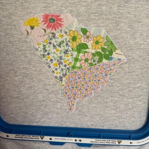K&ouml;nnte beinhalten: Ein graues T-Shirt mit einem floralen Patchwork-Design in Gelb-, Rosa- und Lilat&ouml;nen. Das Design hat die Form eines Staates. Das Shirt befindet sich in einem blauen Stickrahmen.