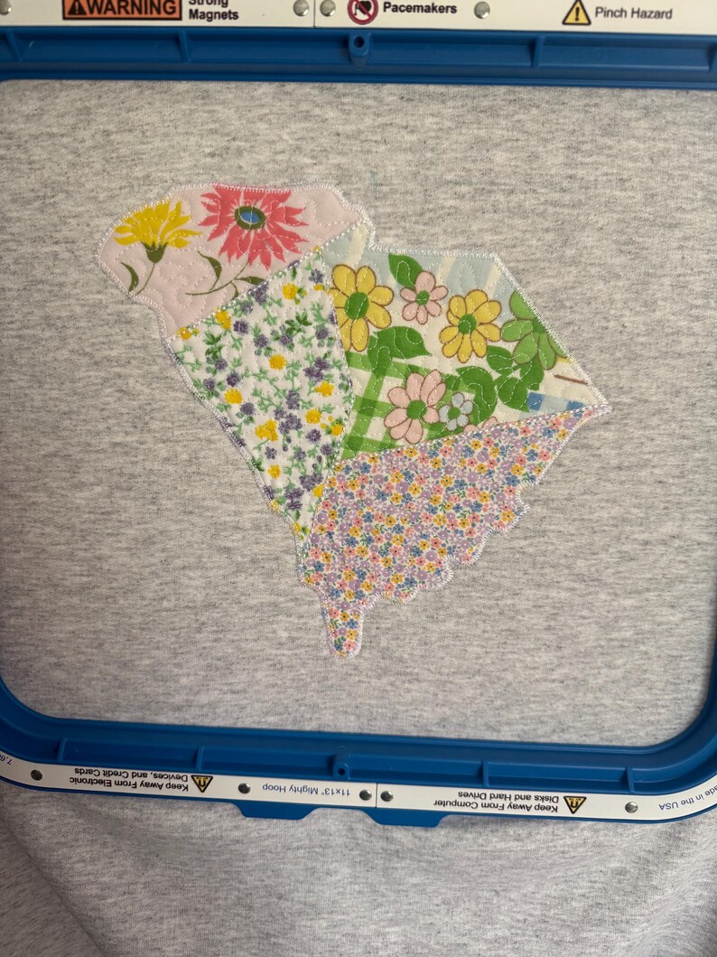 K&ouml;nnte beinhalten: Ein graues T-Shirt mit einem floralen Patchwork-Design in Gelb-, Rosa- und Lilat&ouml;nen. Das Design hat die Form eines Staates. Das Shirt befindet sich in einem blauen Stickrahmen.