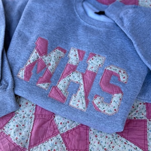 May include: A gray sweatshirt with the letters "NHS" appliqued on the front in pink fabric with a floral pattern. The sweatshirt is lying on a pink and white floral quilt.