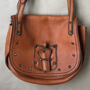 May include: A brown leather satchel bag with a flap closure and a metal buckle. The bag has a long adjustable strap and is decorated with silver studs.