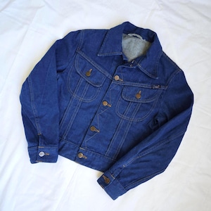 Könnte beinhalten: Eine dunkelblaue Jeansjacke mit spitzem Kragen, langen Ärmeln mit geknöpften Manschetten und einer Knopfleiste vorne. Die Jacke verfügt über zwei Brusttaschen mit geknöpften Klappen und sichtbare kupferfarbene Nähte am gesamten Kleidungsstück.