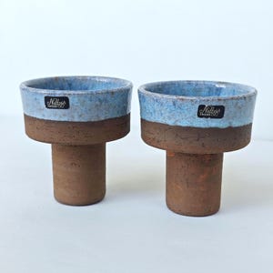 May include: Two ceramic cups with a cylindrical base and a wide, flared top. The lower portion is a textured, matte brown clay, while the upper rim features a speckled, glossy light blue glaze. Each cup has a black label reading Nillsjö Sweden.