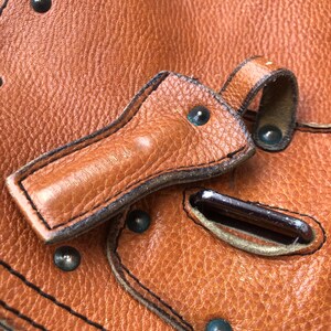 May include: Close-up of a brown leather bag with a flap closure and metal hardware. The bag has a textured surface and is decorated with stitching and rivets.