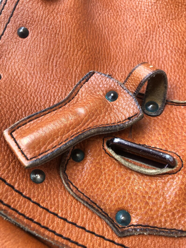 May include: Close-up of a brown leather bag with a flap closure and metal hardware. The bag has a textured surface and is decorated with stitching and rivets.