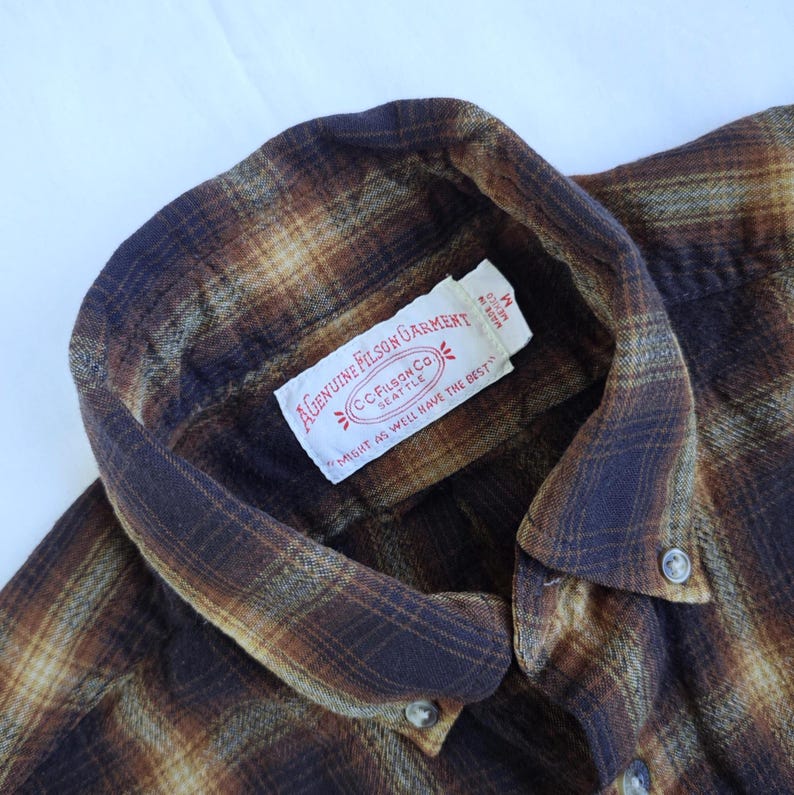 May include: Close-up of a brown and tan plaid flannel shirt. The shirt features a collar, buttons, and a tag that reads "GENUINE FILSON GARMENT" and "MIGHT AS WELL HAVE THE BEST". The shirt is a medium size.