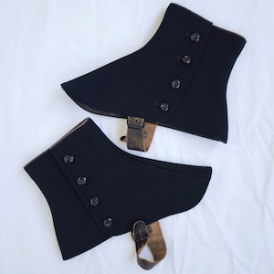 May include: A pair of black spats featuring a curved silhouette with four dark buttons arranged in a vertical row along the side. Each piece includes a brown leather strap with a metal buckle attached at the bottom for securing around the footwear.