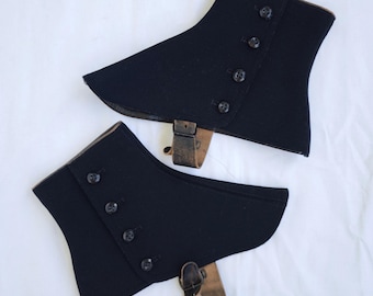 May include: A pair of black spats featuring a curved silhouette with four dark buttons arranged in a vertical row along the side. Each piece includes a brown leather strap with a metal buckle attached at the bottom for securing around the footwear.