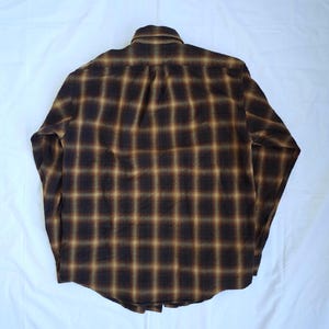 May include: A long-sleeved, button-down shirt with a plaid pattern in shades of brown and tan. The shirt is laid flat, showcasing the back with the collar and button details visible. The fabric appears to be a soft, woven material.
