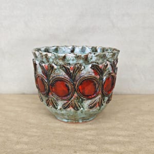 Può includere: Un vaso in ceramica caratterizzato da una smaltatura testurizzata e maculata nei toni del verde e dell'azzurro. L'esterno è decorato con un motivo ripetuto di foglie in rilievo marrone scuro che circondano accenti circolari rosso vivo.