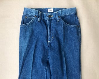 May include: Close-up of blue denim jeans with a classic five-pocket design. The jeans feature a button and zipper closure, belt loops, and the "Lee" brand label. The denim has a medium wash and a slightly worn appearance.