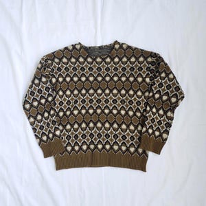 Peut inclure: Un pull en tricot à manches longues présentant un motif géométrique répétitif en forme de losanges dans des tons marron, noir et crème. Le vêtement possède une encolure ronde, des poignets et un ourlet côtelés marron, en maille épaisse.