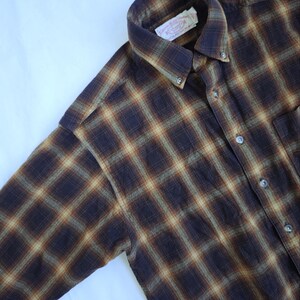 May include: A button-down, long-sleeve shirt with a plaid pattern in shades of brown, tan, and dark blue. The shirt has a button-down collar and a chest pocket. The label on the collar is visible.