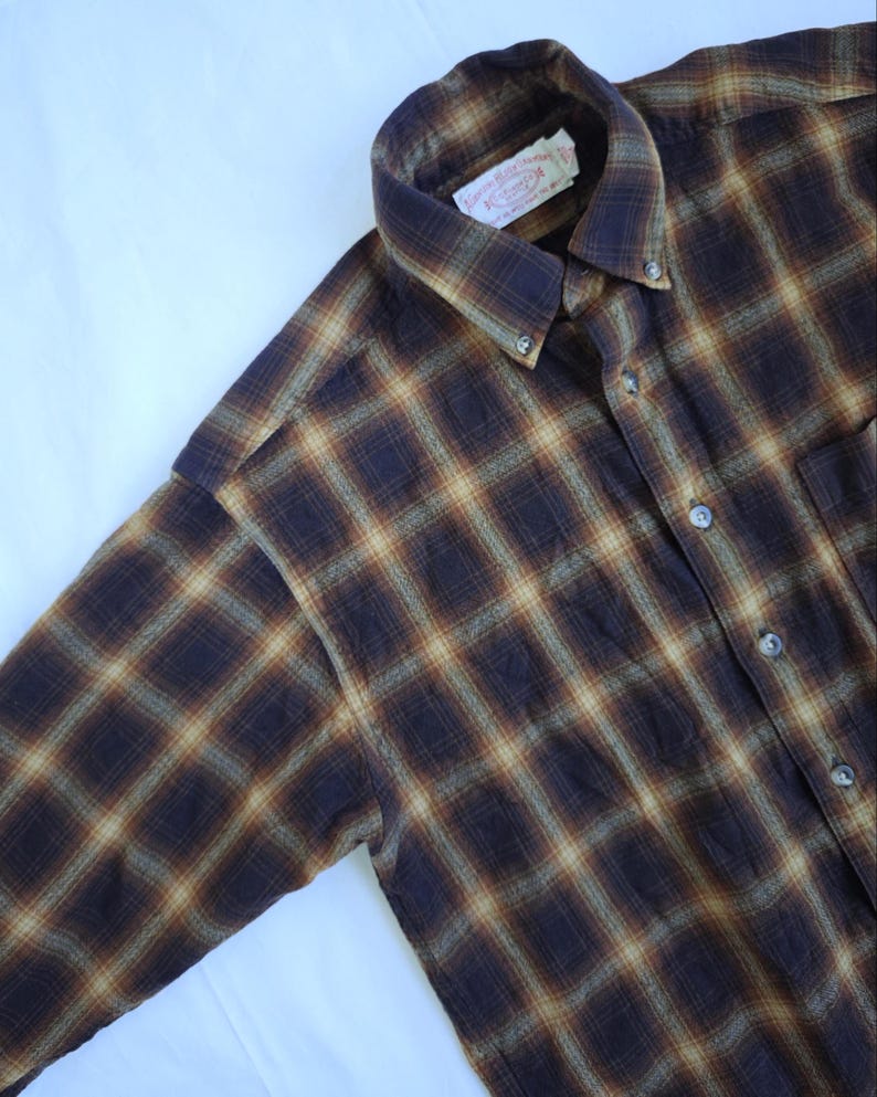 May include: A button-down, long-sleeve shirt with a plaid pattern in shades of brown, tan, and dark blue. The shirt has a button-down collar and a chest pocket. The label on the collar is visible.