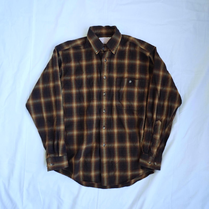 May include: A long-sleeved, button-down shirt in a brown and tan plaid pattern. The shirt features a button-down collar, a single chest pocket, and buttoned cuffs. The fabric appears to be a soft, flannel-like material.