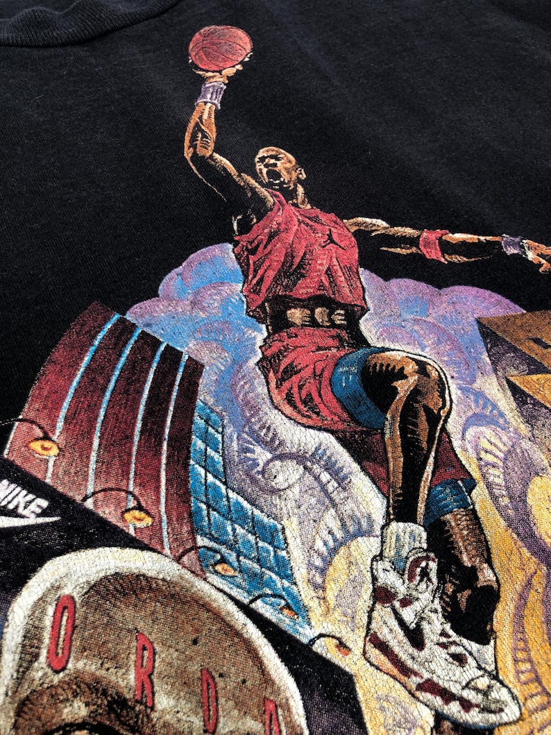 Może przedstawiać: Czarny T-shirt z nadrukiem graficznym koszykarza w powietrzu, ubranym w czerwoną koszulkę i białe buty sportowe, z widocznym napisem "NIKE" w lewym dolnym rogu koszulki.