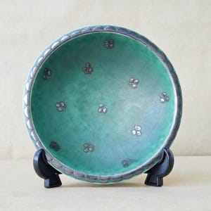 Può includere: Una ciotola rotonda in ceramica verde caratterizzata da una sottile finitura smaltata a craquelé e da una serie di piccoli motivi floreali color argento sparsi sulla superficie interna. Il bordo è decorato con un motivo a festoni in argento.