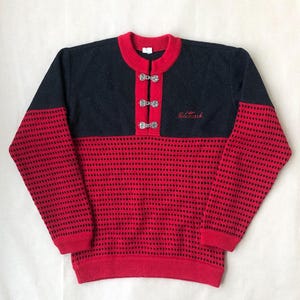 May include: A red and black knitted sweater with a high neckline and silver buttons. The sweater has a "Telemark" logo embroidered on the front.