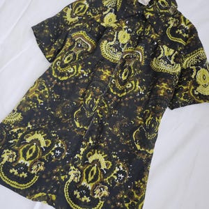 Può includere: Camicia a maniche corte con motivo paisley giallo, marrone e bianco su sfondo scuro. La camicia ha un colletto e una chiusura con bottoni. Il tessuto sembra leggero.