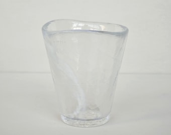 Op de afbeelding: Een doorzichtig glas met een onregelmatige, organische vorm en een licht uitlopende rand. Het glas bevat subtiele, wervelende witte insluitingen in het transparante materiaal, wat zorgt voor een getextureerd uiterlijk over het hele lichaam van de beker.