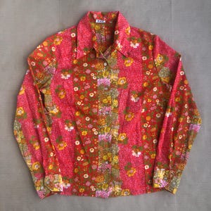 Può includere: Una camicia a maniche lunghe con stampa floreale rossa e gialla con colletto abbottonato. La camicia ha un'etichetta 'WAG' sul colletto.