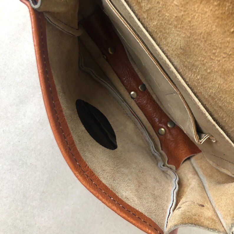 May include: The inside of a brown leather bag with a flap closure. The bag has a small pocket with a black fabric lining. The bag is lined with a light brown fabric.