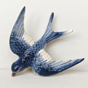 May include: A ceramic wall ornament shaped like a flying swallow bird. The bird features a glossy finish with deep blue and white plumage, a pointed tan beak, and a single visible small red eye. The wings are extended in a dynamic flight pose.