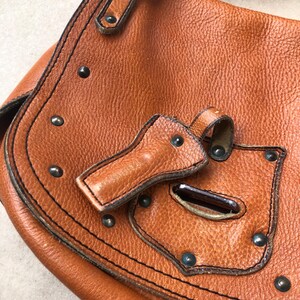 May include: A brown leather satchel bag with a flap closure and metal studs. The bag has a small pocket on the front with a metal buckle.