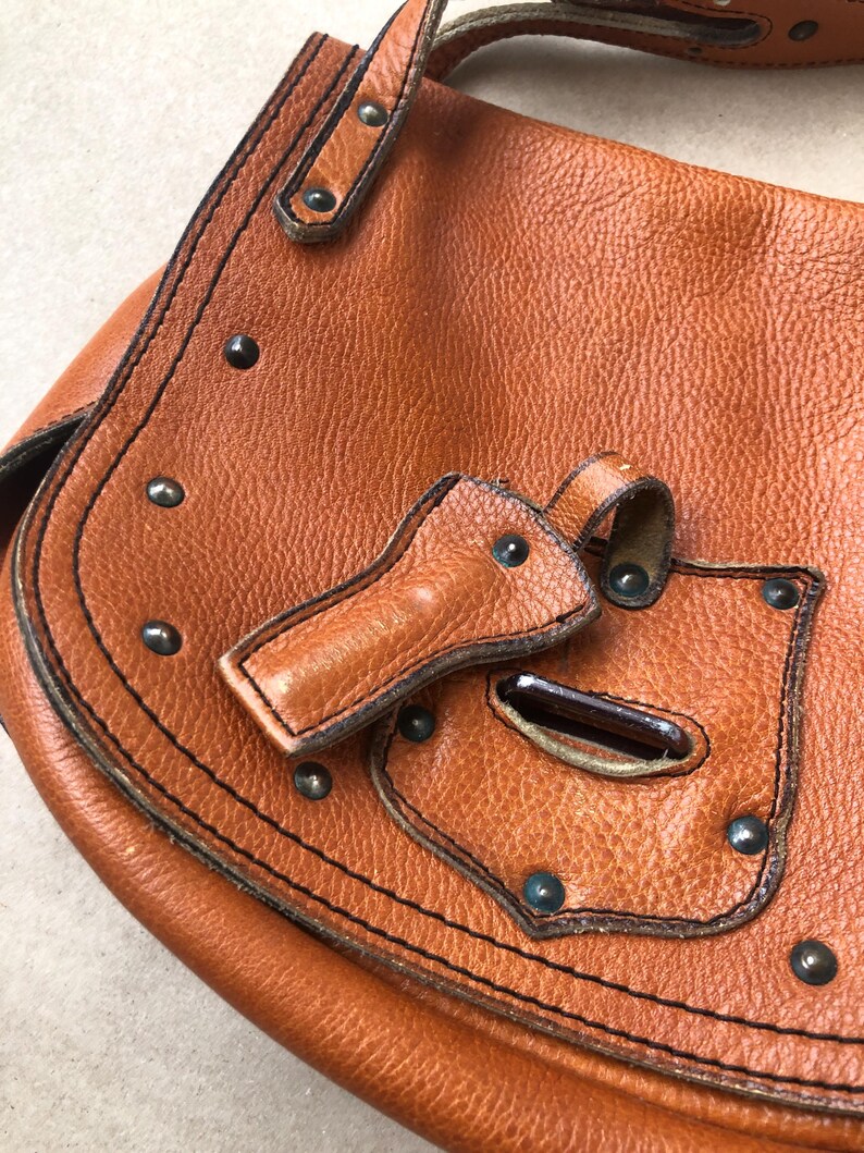 May include: A brown leather satchel bag with a flap closure and metal studs. The bag has a small pocket on the front with a metal buckle.