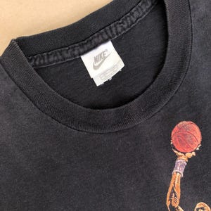 Może przedstawiać: Czarny T-shirt z grafiką koszykarza i metką z logo Nike. Koszykarz ma na sobie czerwoną i czarną koszulkę i trzyma piłkę do koszyk&oacute;wki.