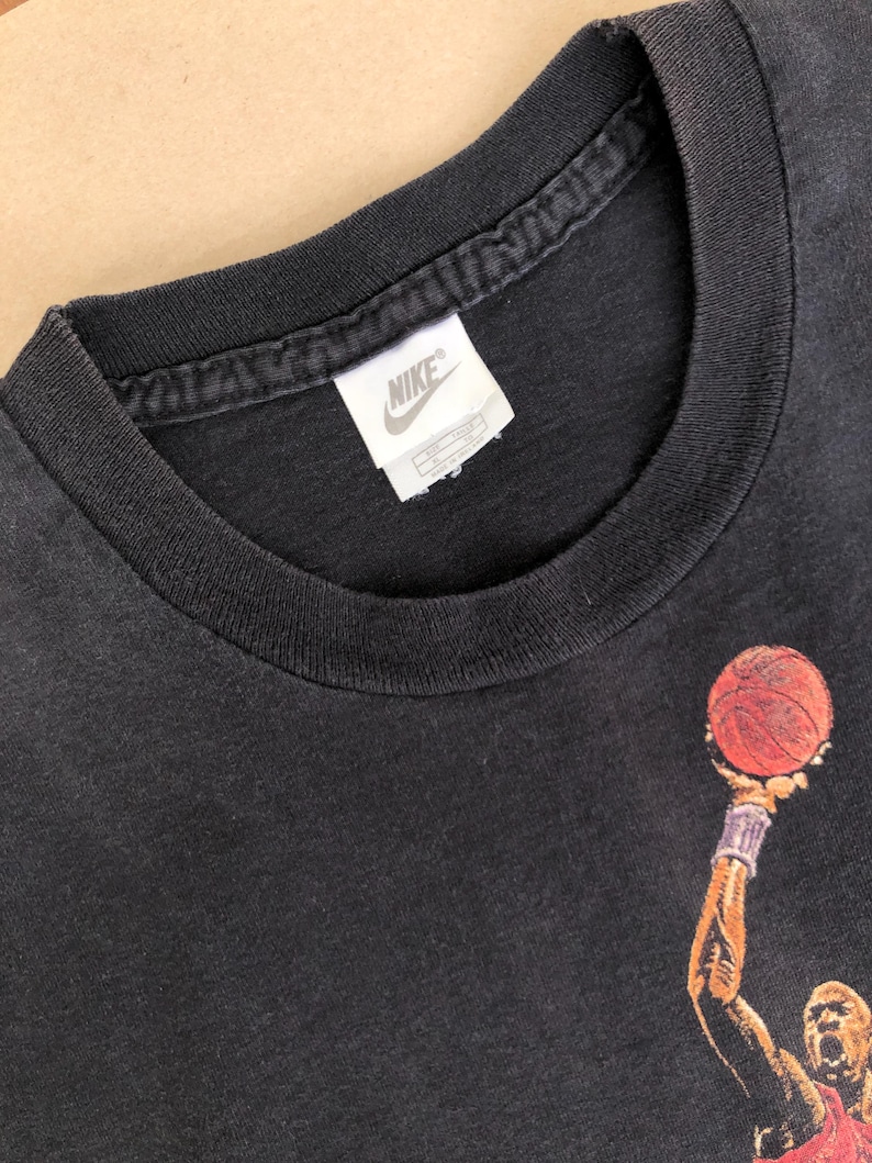 Może przedstawiać: Czarny T-shirt z grafiką koszykarza i metką z logo Nike. Koszykarz ma na sobie czerwoną i czarną koszulkę i trzyma piłkę do koszyk&oacute;wki.