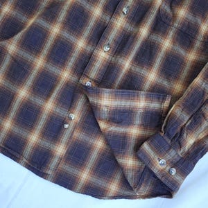 May include: A button-down plaid shirt in shades of brown, tan, and navy blue. The shirt features a classic plaid pattern, a button-down front, a chest pocket, and buttoned cuffs. The fabric appears to be a soft, woven material.