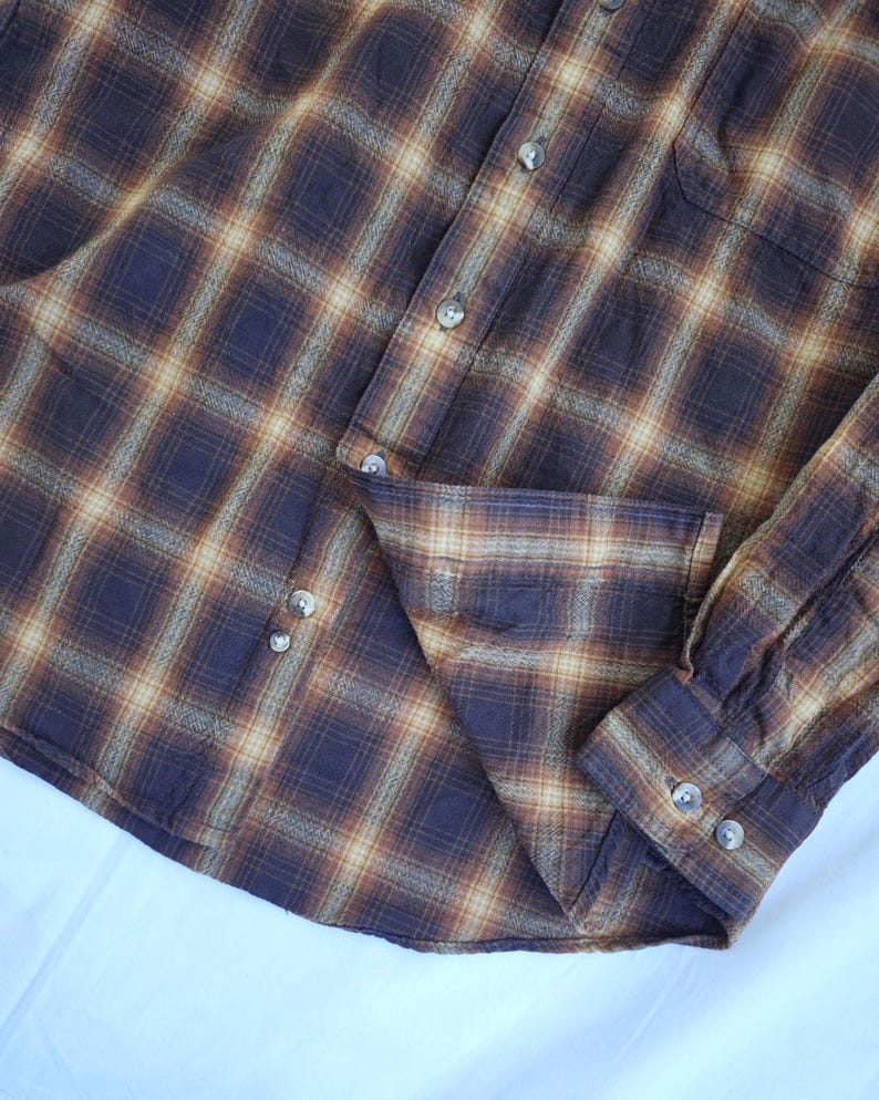 May include: A button-down plaid shirt in shades of brown, tan, and navy blue. The shirt features a classic plaid pattern, a button-down front, a chest pocket, and buttoned cuffs. The fabric appears to be a soft, woven material.