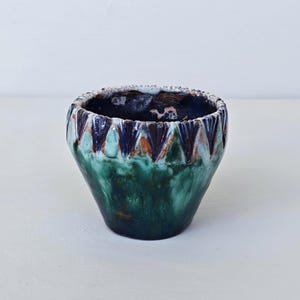 Puede incluir: Un cuenco de cerámica con una base de color verde oscuro que transiciona a un esmalte turquesa claro y marrón cerca del borde. El borde superior está festoneado con una serie de hendiduras verticales y el interior tiene un acabado brillante.