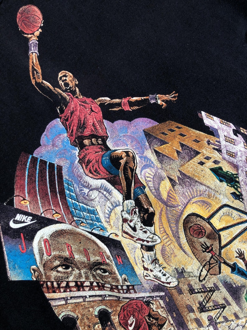 Może przedstawiać: Czarny vintage T-shirt z nadrukiem graficznym koszykarza w powietrzu, wykonującego wsad. Koszykarz ma na sobie czerwoną i białą koszulkę oraz białe buty. Tło to kolorowe miasto z budynkami i chmurami.