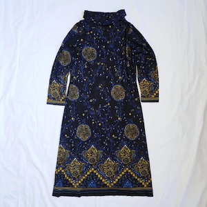 Könnte beinhalten: Ein langärmeliges Kleid mit schwarzer Grundfarbe und einem sich wiederholenden Muster aus goldenen und blauen Blumenmotiven, geschwungenen Ranken und geometrischen Bordüren. Das Kleid hat einen hohen Ausschnitt und eine gerade Passform.
