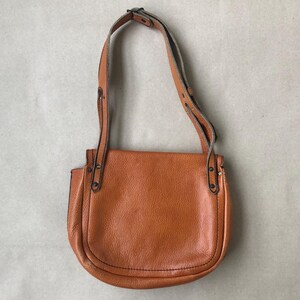 May include: A brown leather satchel bag with a long adjustable strap. The bag has a flap closure and a rounded shape.