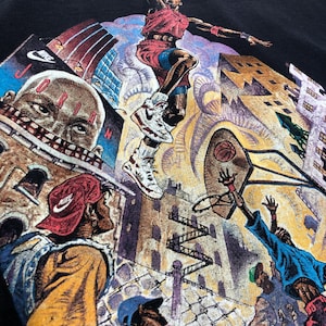 Może przedstawiać: Czarny vintage'owy t-shirt z kolorowym nadrukiem graficznym przedstawiającym koszykarza w powietrzu, otoczonego budynkami i innymi ludźmi. Koszykarz ma na sobie czerwoną i białą koszulkę oraz czarne spodenki. Na koszulce widoczny jest napis "NIKE" i "JORDAN".