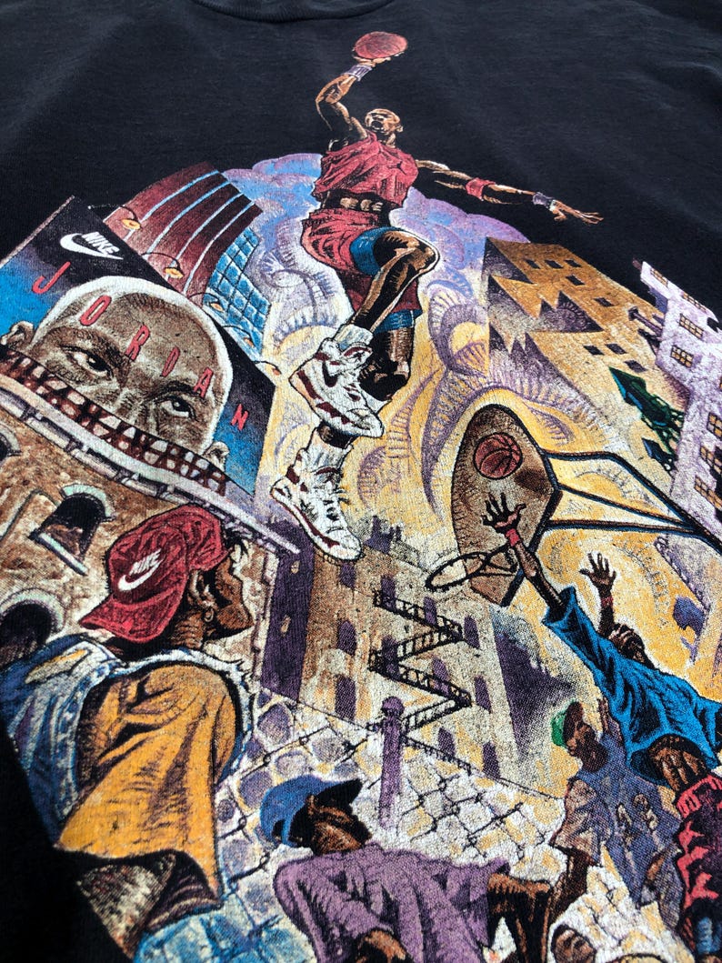 Może przedstawiać: Czarny vintage'owy t-shirt z kolorowym nadrukiem graficznym przedstawiającym koszykarza w powietrzu, otoczonego budynkami i innymi ludźmi. Koszykarz ma na sobie czerwoną i białą koszulkę oraz czarne spodenki. Na koszulce widoczny jest napis "NIKE" i "JORDAN".