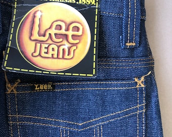 Puede incluir: Primer plano de unos vaqueros de mezclilla azul oscuro con un bolsillo trasero y costuras amarillas. Una etiqueta dice "Lee Jeans" y "Founded Kansas, 1889." Los vaqueros miden 66 x 81 cm y son 100% algodón.