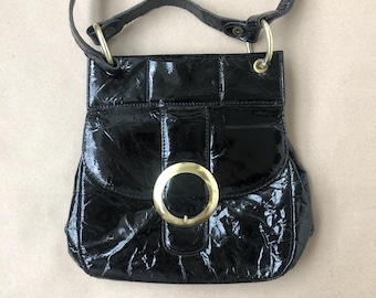 May include: A black leather crossbody bag with a gold buckle and a circular design. The bag has a textured, crinkled look.