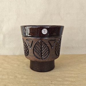 May include: A dark brown ceramic planter with a glossy finish on the rim and a matte, textured base. The exterior features embossed leaf and triangle patterns around the center. A small white circular sticker reads Hand Made In Sweden.