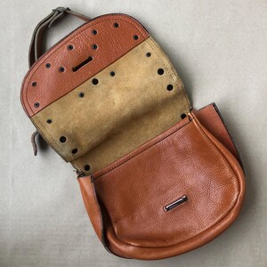May include: A brown leather satchel bag with a flap closure and a metal buckle. The bag has a suede lining and a brown leather strap.