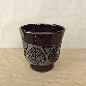 May include: A dark brown ceramic cup with a glossy finish on the rim and base. The middle section features a textured, matte brown band decorated with a repeating pattern of embossed leaf shapes and triangles encircling the entire vessel.