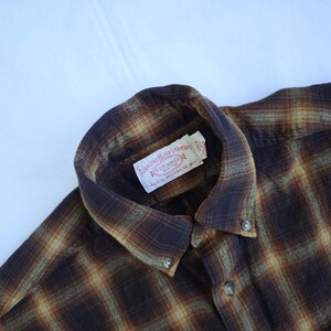 May include: A brown and tan plaid flannel shirt with a button-down collar. The shirt has a label that reads "GENUINE FILSON GARMENT" and "CC FILSON CO. SEATTLE". The shirt is made of a soft, textured fabric.