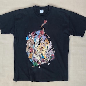 Może przedstawiać: Czarny T-shirt z nadrukiem graficznym koszykarza w czerwonym stroju, otoczonego innymi postaciami i budynkami. Grafika jest w stylu komiksowym.