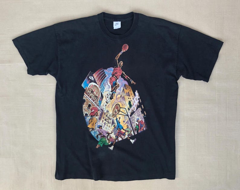 Może przedstawiać: Czarny T-shirt z nadrukiem graficznym koszykarza w czerwonym stroju, otoczonego innymi postaciami i budynkami. Grafika jest w stylu komiksowym.